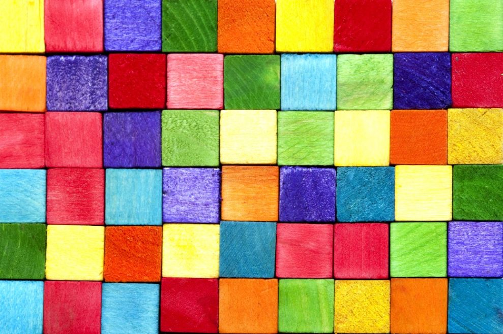 Bloquinhos quadrados de madeira empilhados um em cima do outro fazendo uma parede. Tem blocos de várias cores, azul marinho, azul claro, vermelho, rosa, laranja, amarelo, verde escuro, verde claro e vinho