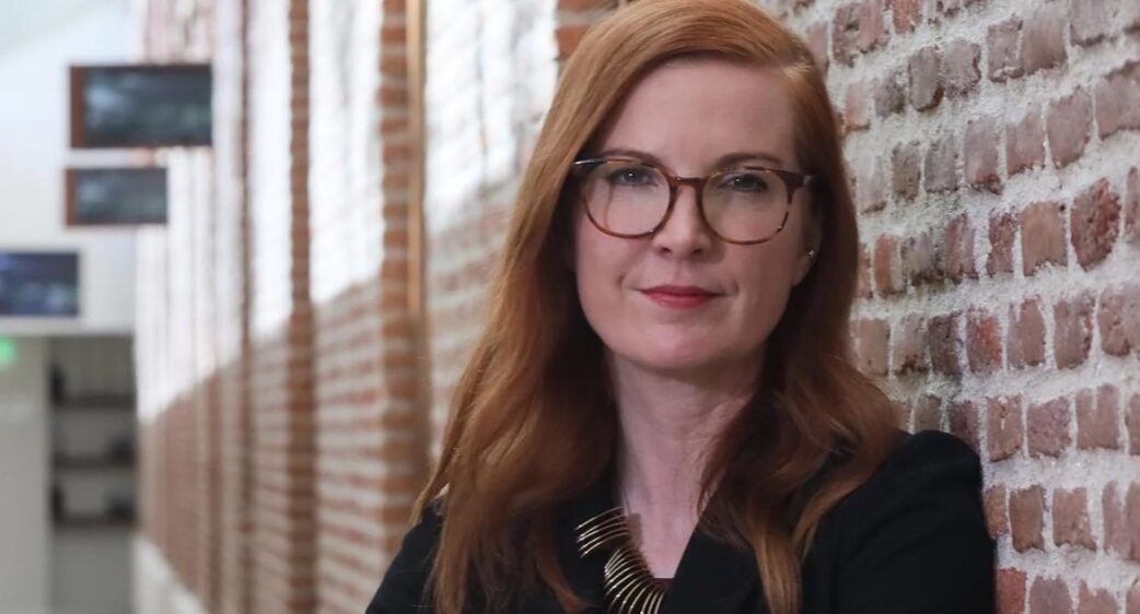 Kate Crawford, cofundadora do AI Now Institute de óculos e cabelos ruivos compridos, encostada em uma parede de tijolos aparente