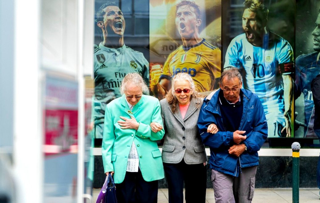 Duas senhoras e um senhor estão de braços dados, cabeça baixa e sorrindo. Está frio, pois os três estão de casaco. Ao fundo um painel grande mostrando fotos dos jogadores Cristiano Ronaldo, Messi, Neymar como se estivessem comemorando um gol.