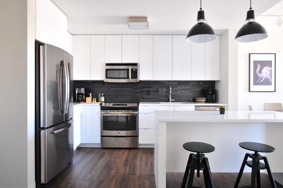 Cozinha super moderna e arrumada. Armários em branco, eletrodomésticos metalizados, banquetas e luminárias pretas. A geladeira grande, com freezer na parte de cima fica do lado esquerdo da foto. Do lado direito tem uma bancada branca estilo americano com 2 banquetas pretas. Ao fundo, os armários com o fogão e o micro-ondas logo acima dele. Tem um quadro de pássaro estilizado do lado direito na parede atrás da bancada. Tem alguns objetos em cima da bancada do fogão em todos que combinam com o resto das cores da cozinha.