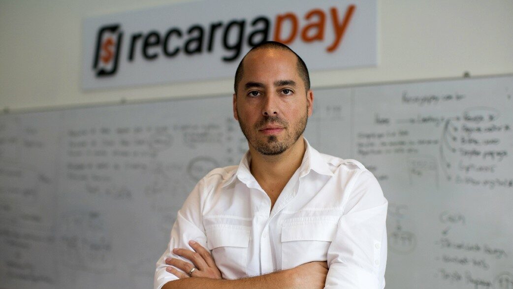 A foto mostra o empreendedor Rodrigo Teijeiro, fundador da fintech recargapay. Ele está de pé, com braços cruzados, usa uma camisa branca. Atrás dele, uma lousa com gráficos e textos e o logotipo da empresa