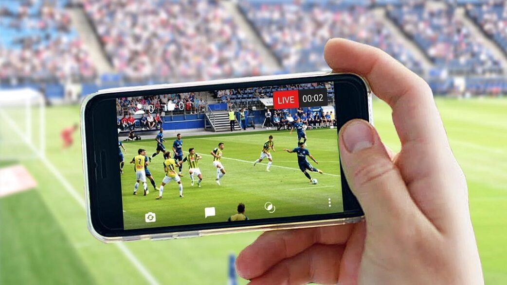 Torcedor de futebol assiste a uma partida em um estádio enquanto segura um smartphone que transmite uma partida de futebol