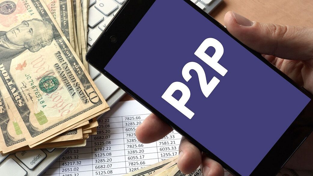 Com notas de dólar ao fundo, uma mão segura um celular em cuja tela está escrito "P2P"