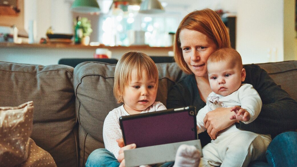 Mãe segura dois filhos pequenos em um sofá dentro de uma casa. A criança mais velha carrega um tablet nas mãos.