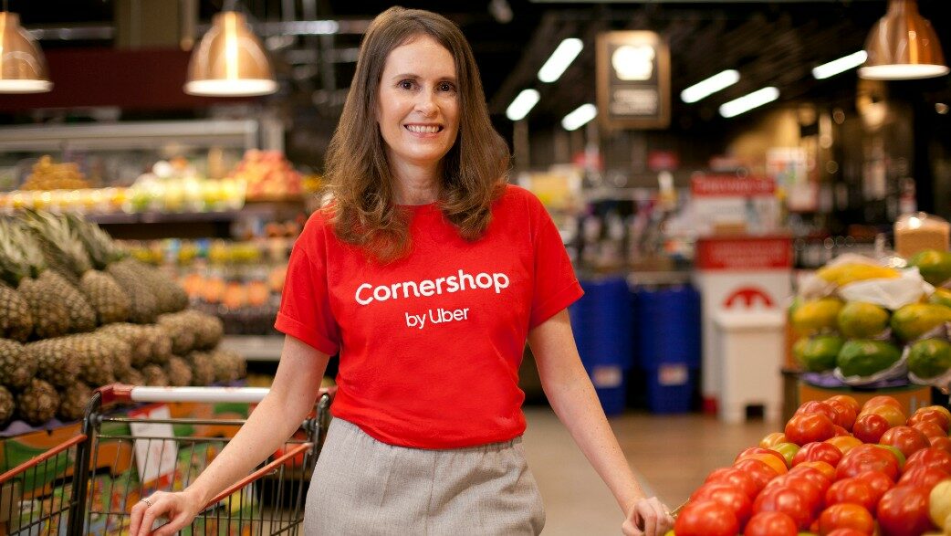 Cristina Alvarenga, head da Cornershop no Brasil, está posando sorrindo, com uma camiseta vermelha, tendo ao fundo as prateleiras e bancadas de frutas de um supermercado