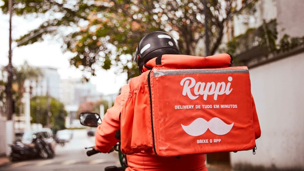 Imagem mostra entregador pilotando uma moto e levando uma caixa de entregas laranja da Rappi nas costas