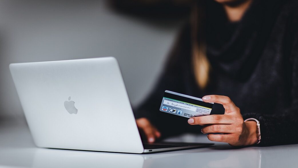 Imagem mostra uma mulher segurando um cartão de crédito prateado em frente a um computador prata a Apple