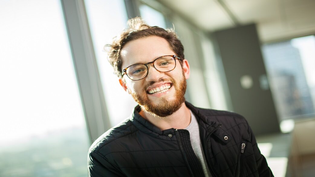 Imagem mostra um homem de cabelo loiro e curto, usando um casaco preto e posando com um sorriso em frente a uma janela