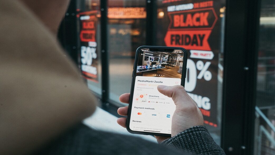 Uma pessoa segura um celular aberto na página de um estabelecimento. A pessoa está parada em frente a uma vitrine com anúncios de descontos da Black Friday