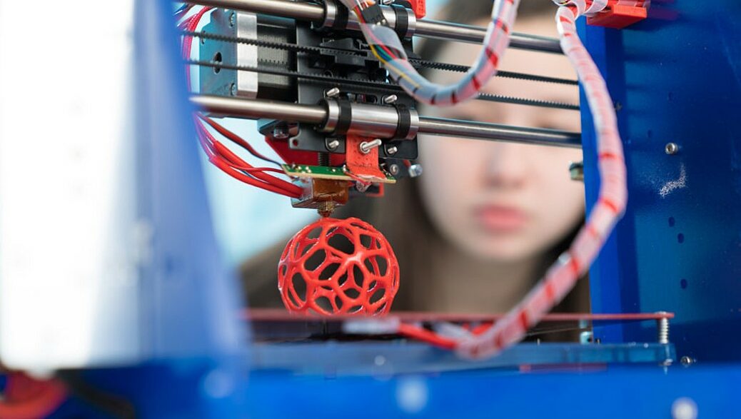 impressora 3D constrói um peça em polímero vermelho, enquanto a pesquisadora observa ao fundo