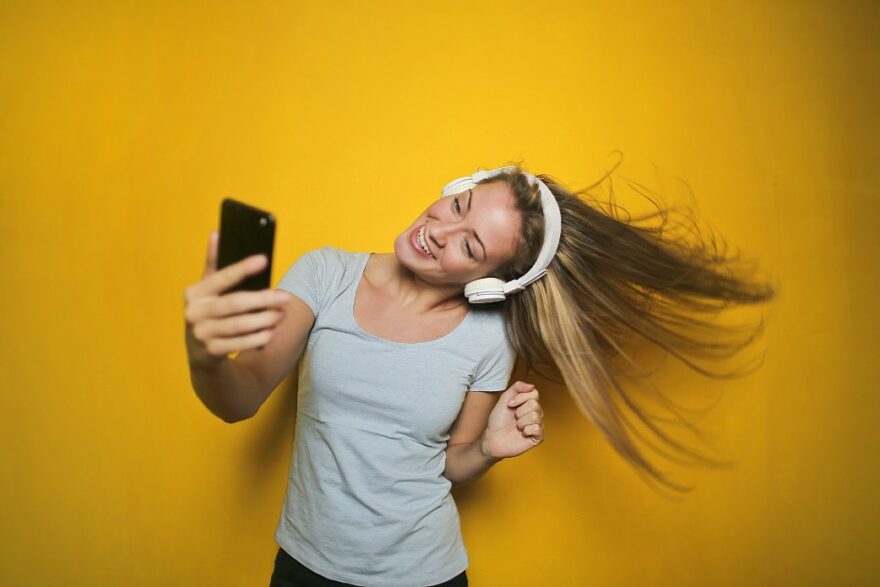 mulher loira de cabelos longos balança a cabeça enquanto ouve música com celular na mão e fones nos ouvidos