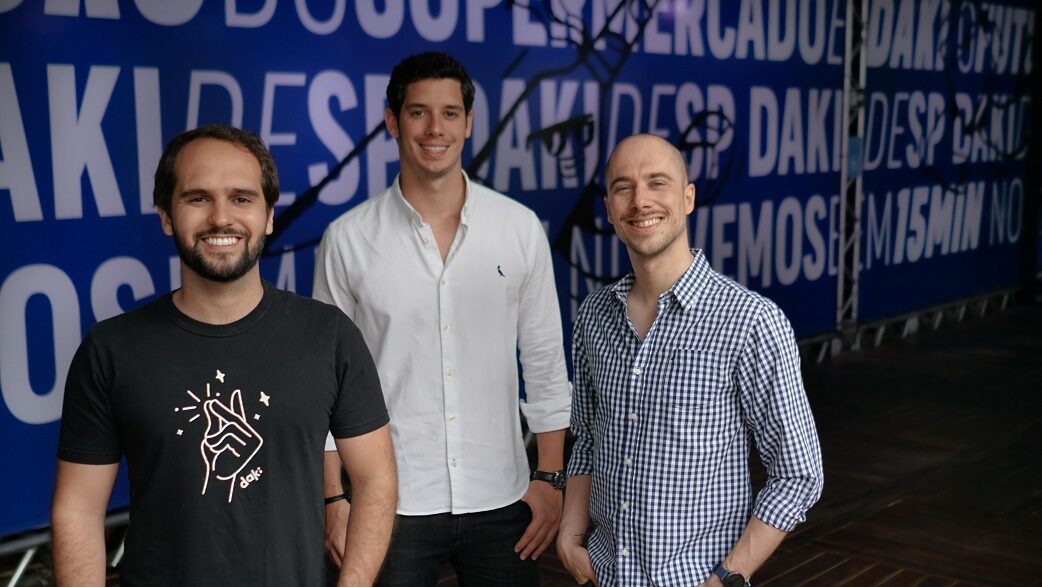 Imagem mostra três homens brancos de pé em frente a uma parede azul