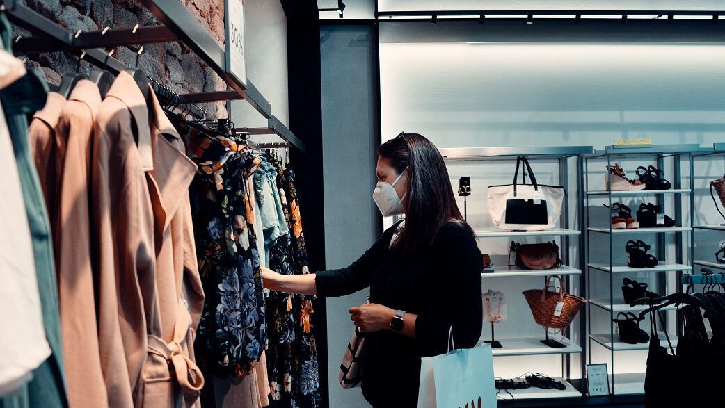 Imagem mostra uma mulher de máscara branca e blusa preta de manga comprida olhando peças de roupa em uma arara de uma loja