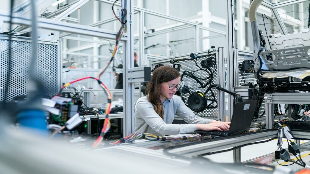 Imagem mostra uma mulher de cabelos longos usando um computador um uma indústria