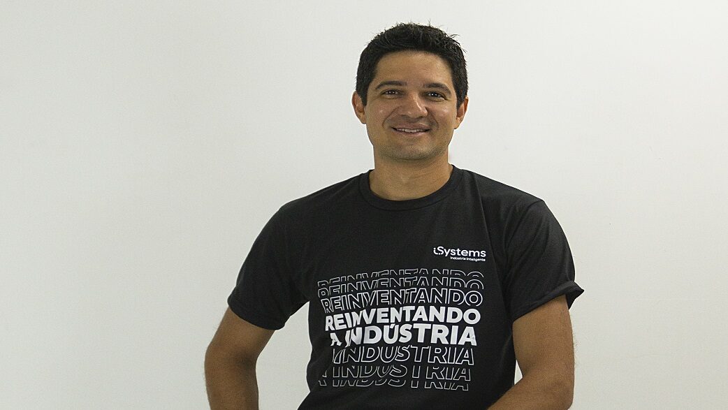 Imagem mostra um homem branco de camiseta preta com os dizeres reinventando a indústria em frente a um fundo branco