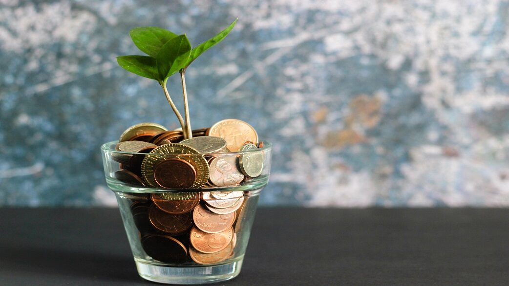 Imagem mostra um vaso de vidro transparente com algumas moedas dentro, das quais surge uma planta