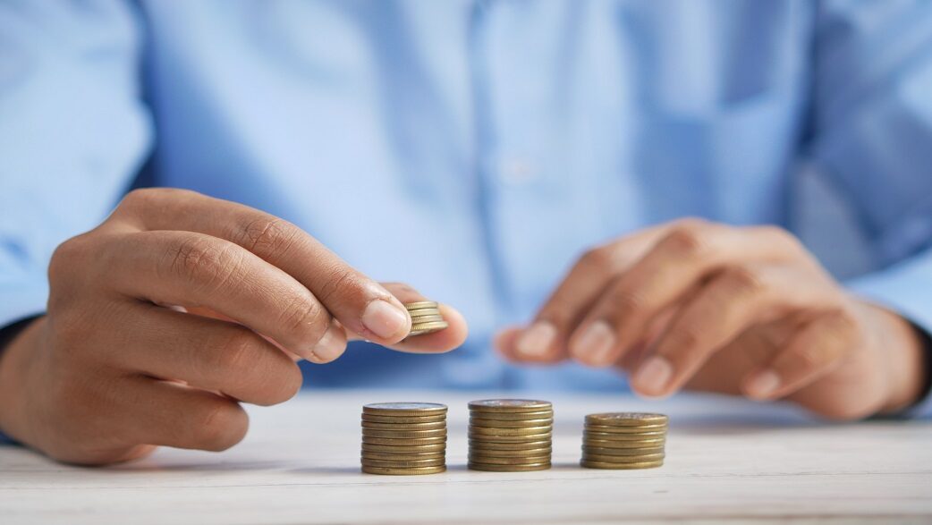 Imagem mostra uma mão colocando uma moeda em uma pilha de moedas