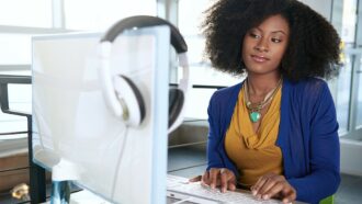 mulher negra de blusa azul olha para tela do computador branco