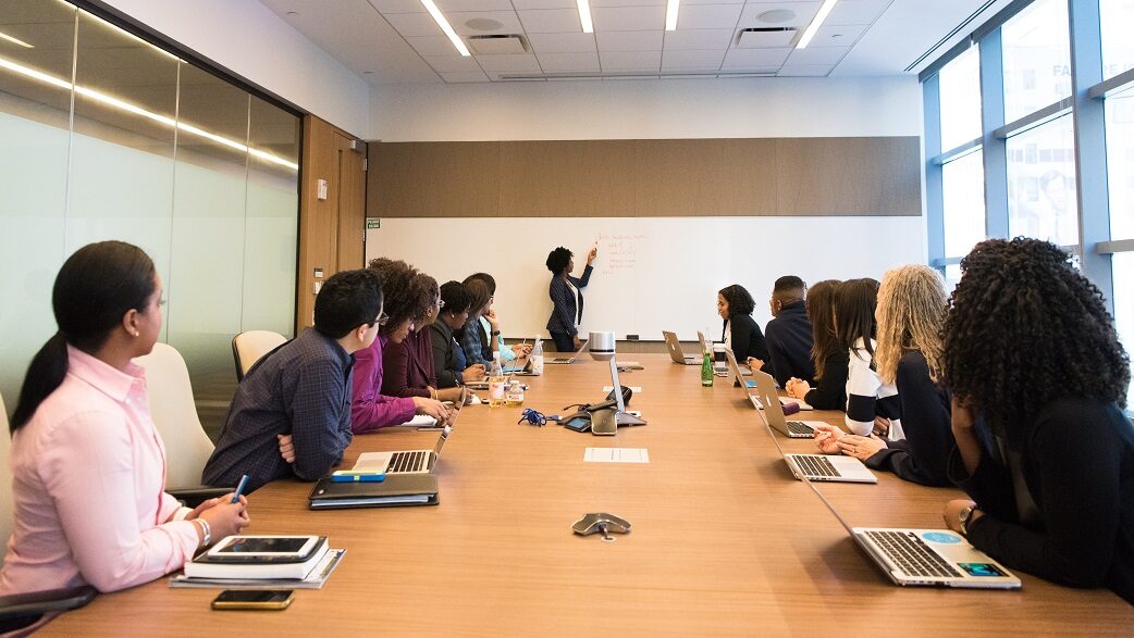 Imagem mostra uma sala de reuniões com uma mesa no centro. Diversas pessoas acompanham uma apresentação.